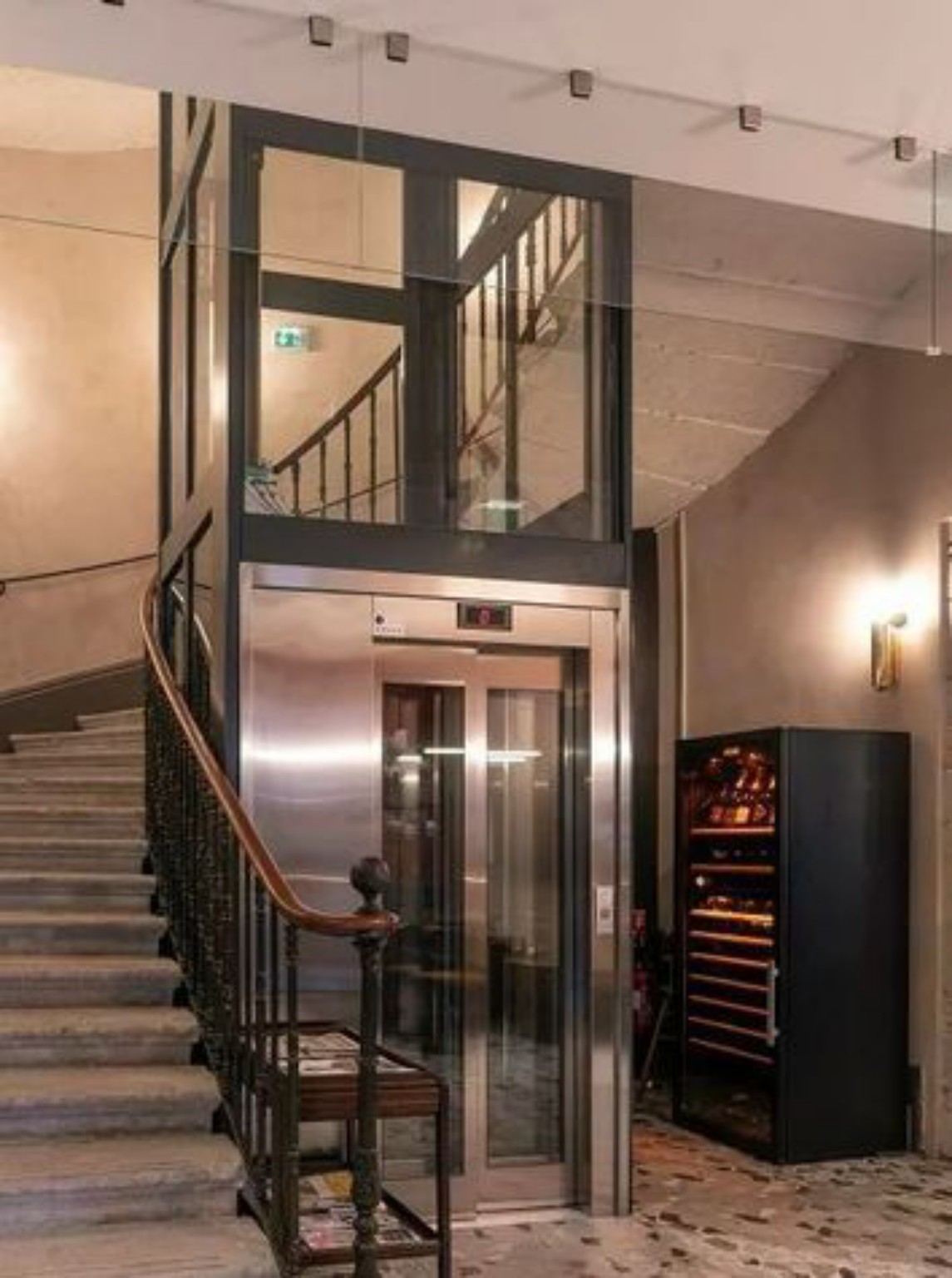 Ascenseur cylindrique en verre et inox installé dans un hall à escalier en fer forgé